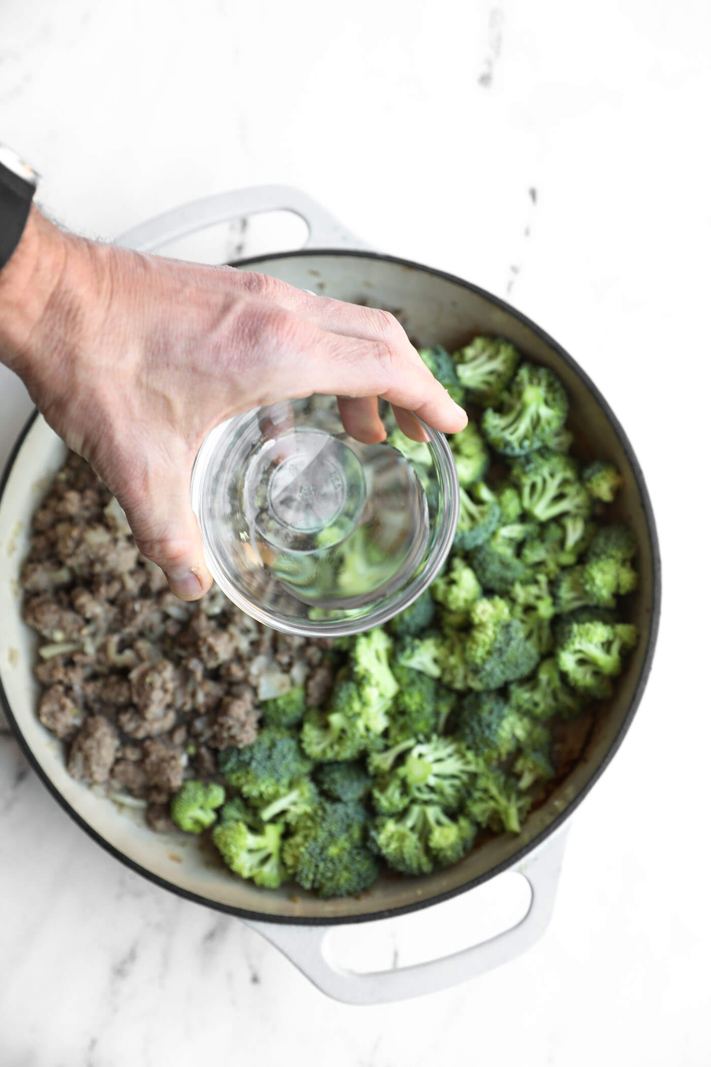 A hand pours water into a pot containing cooked ground meat and fresh broccoli florets.