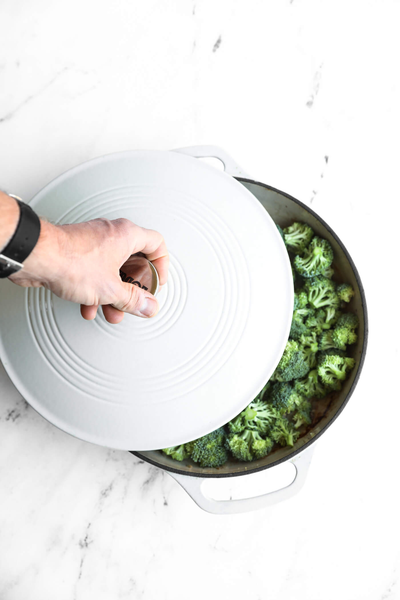 A hand lifts the lid of a white Dutch oven revealing chopped broccoli inside on a white marble surface.