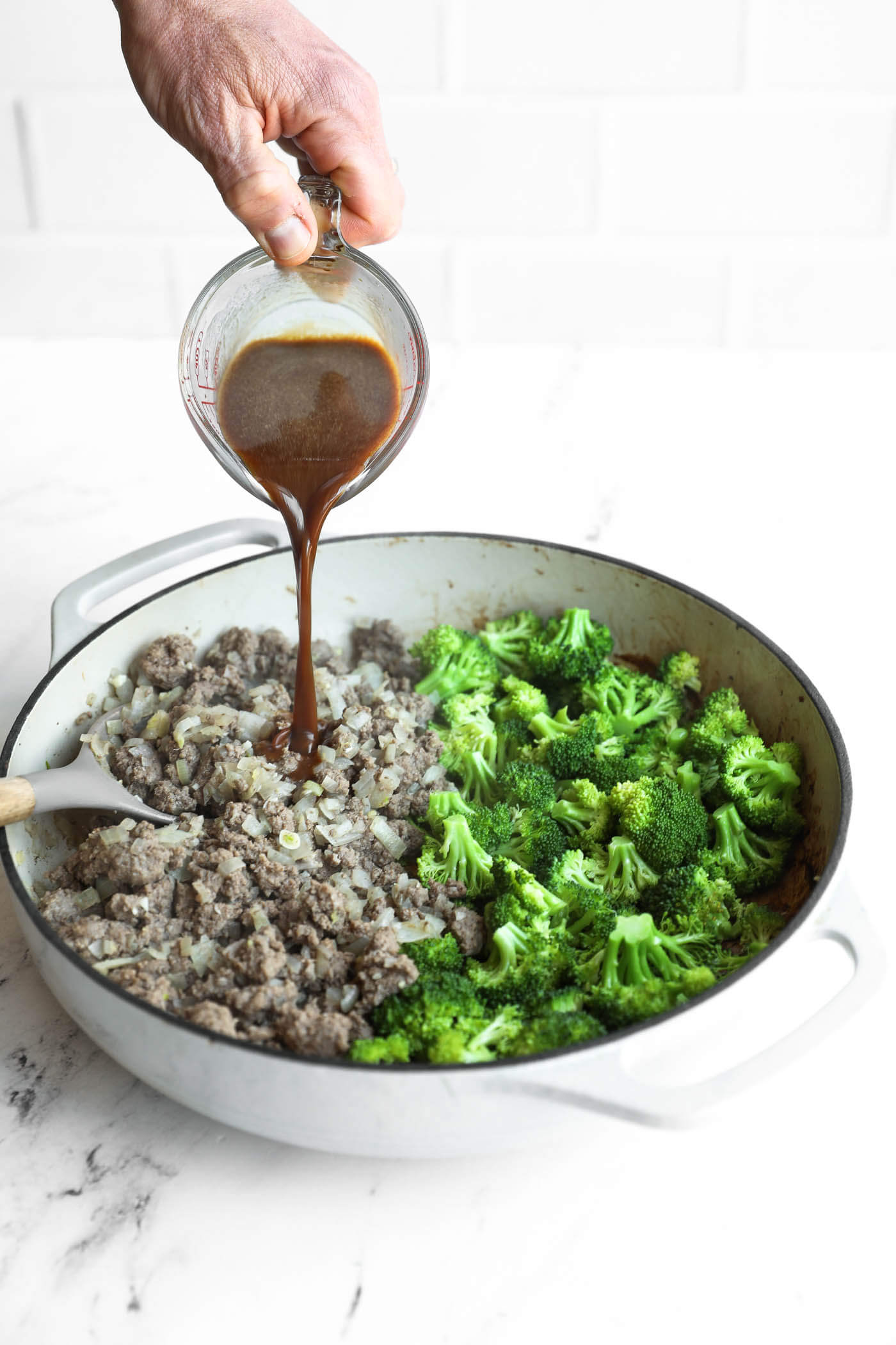 A hand pours brown sauce from a measuring cup into a skillet containing cooked ground meat, onions, and broccoli florets.