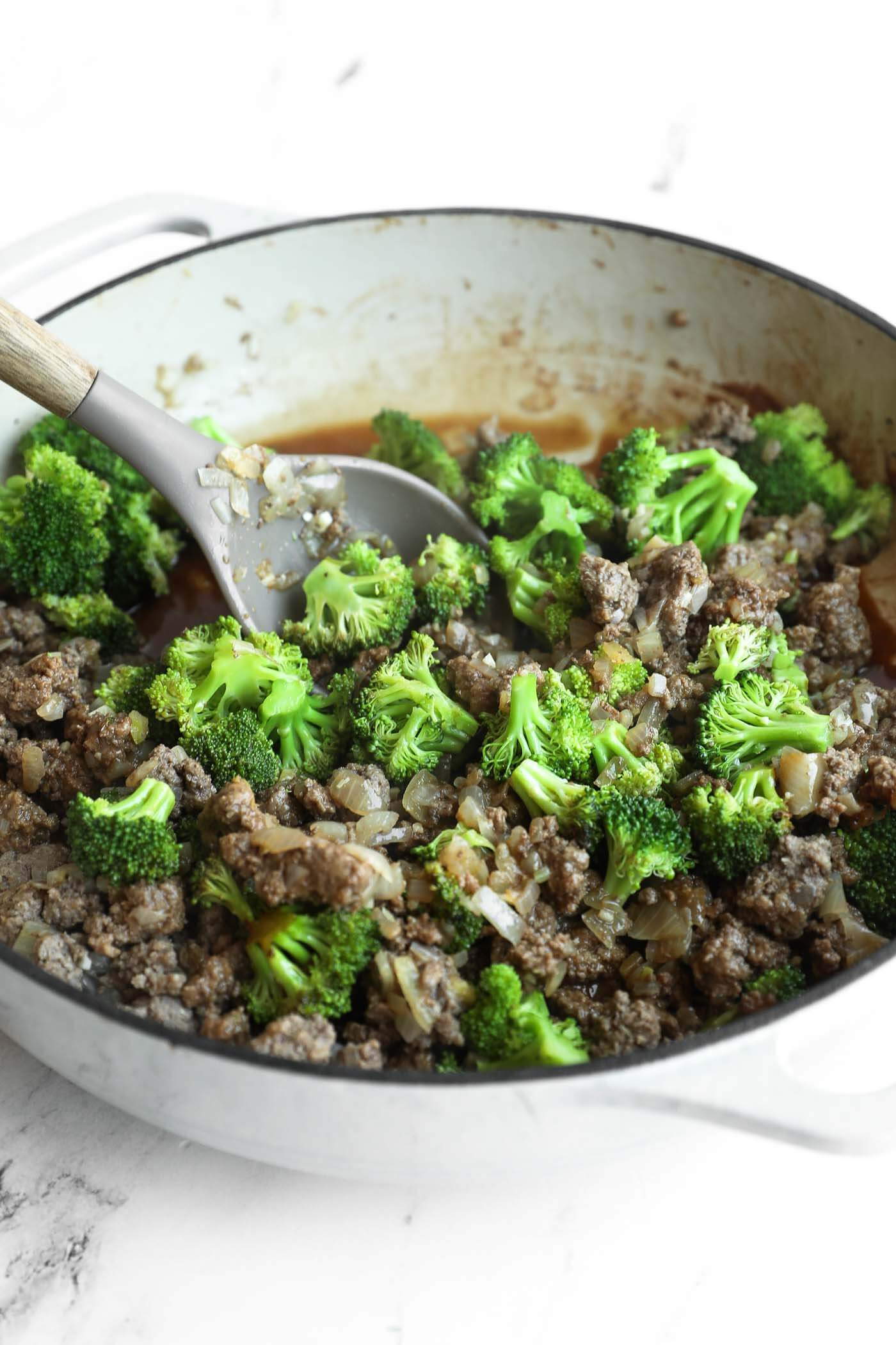 A white pot filled with cooked ground beef, chopped onions, and broccoli florets, with a spoon resting inside.