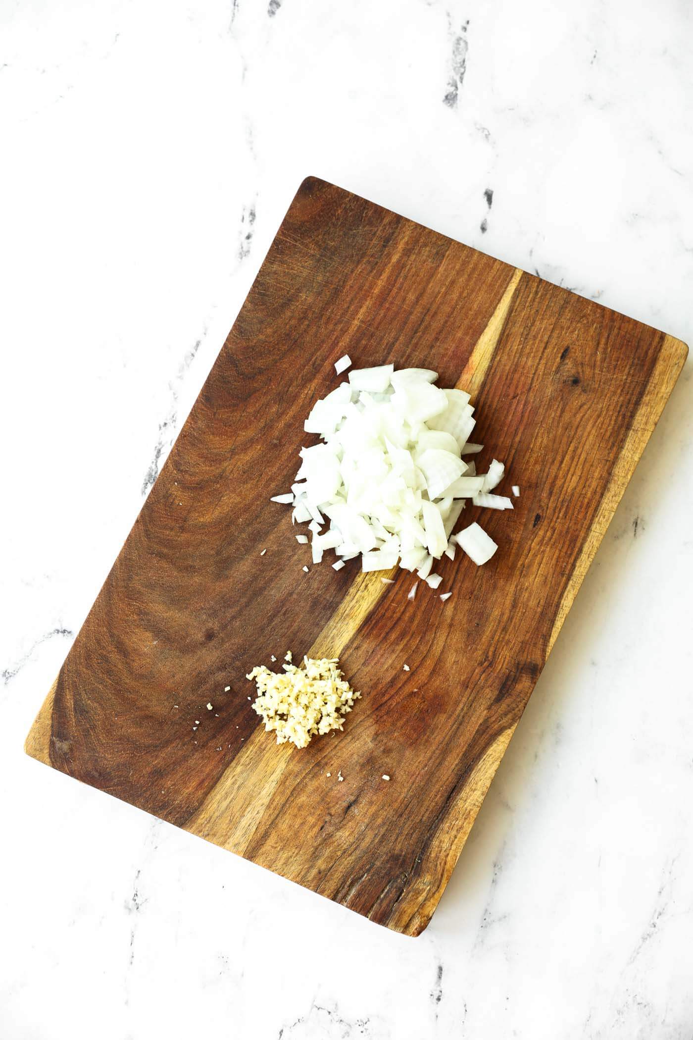 Chopped white onion and minced garlic on a wooden cutting board placed on a white marble surface.