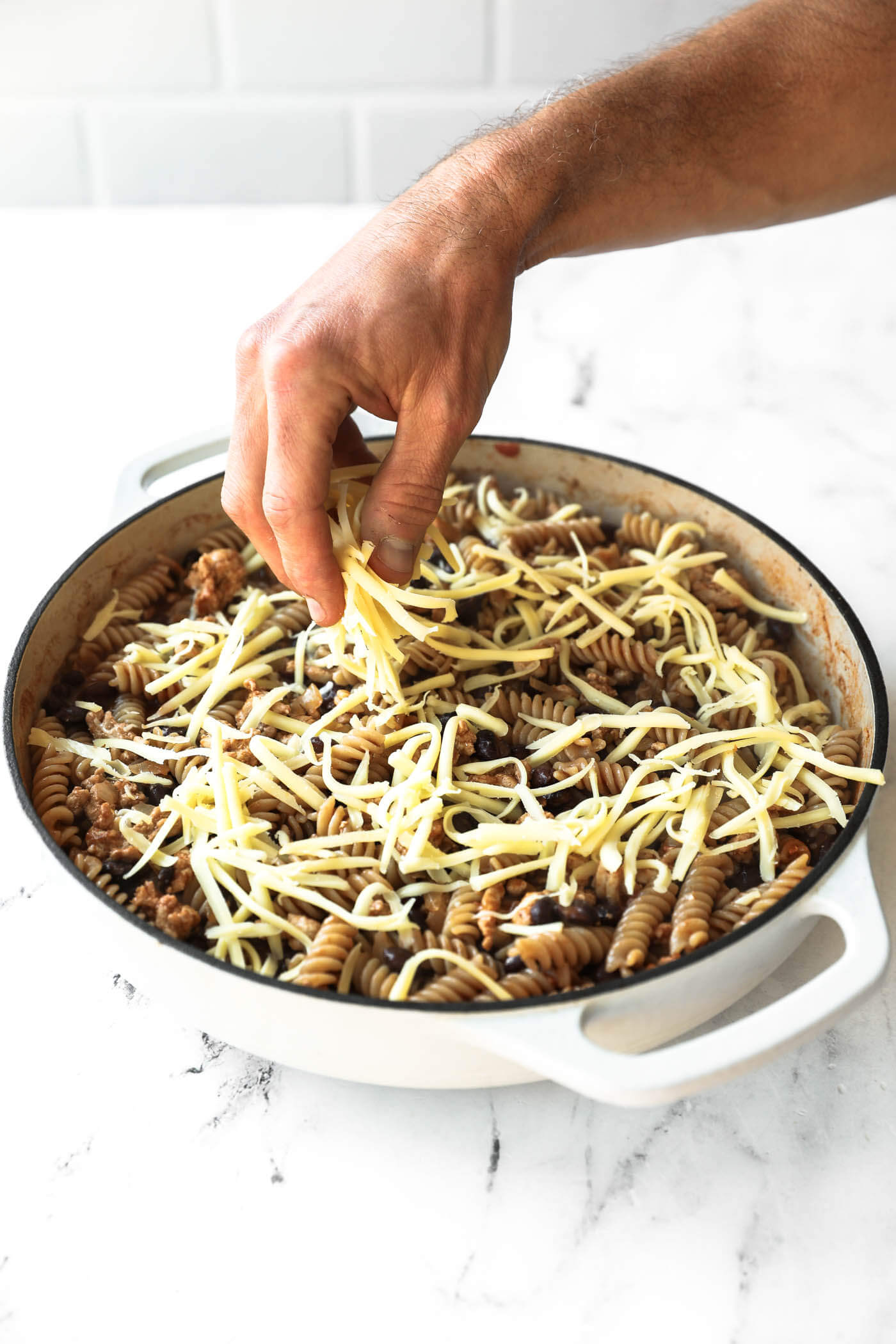 A hand sprinkles shredded cheese over a pan of cooked rotini pasta mixed with ground meat on a white countertop.