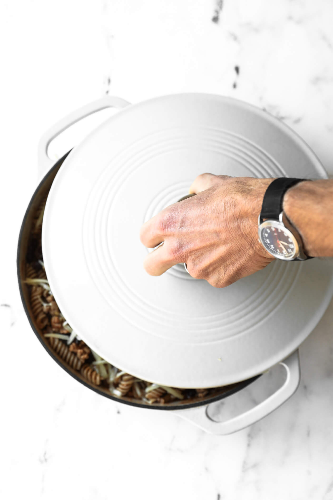 A person wearing a wristwatch lifts the lid off a round, white Dutch oven filled with pasta on a white marble surface.