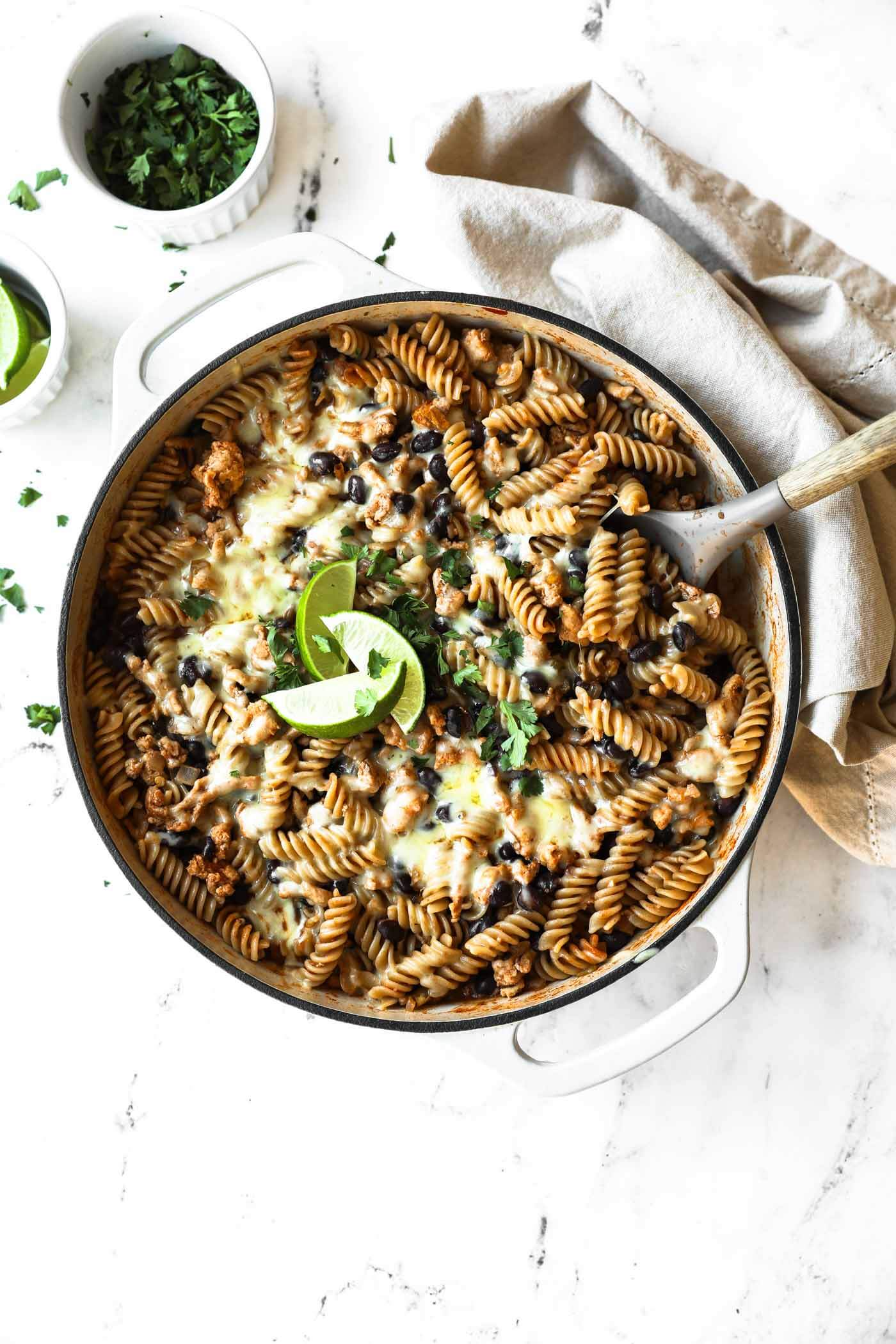 A white pot filled with cheesy rotini pasta bake topped with black beans, lime slices, and fresh herbs. A serving spoon rests in the pot; bowls of herbs are nearby.