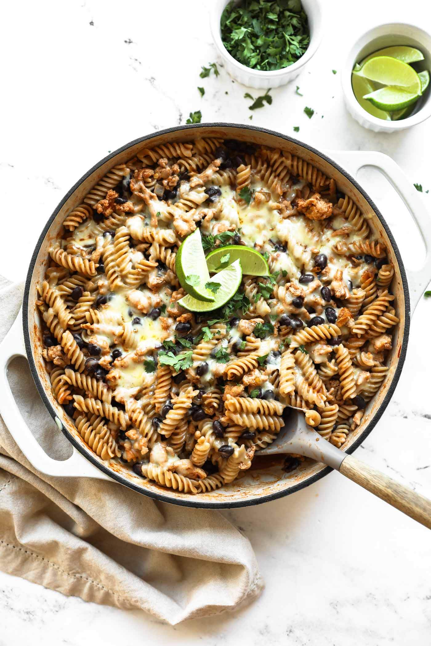 A skillet with cooked rotini pasta, black beans, cheese, ground meat, and fresh cilantro, garnished with lime wedges, on a white surface.