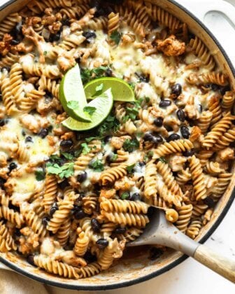 A skillet filled with rotini pasta, black beans, melted cheese, and ground meat, garnished with cilantro and lime wedges, with a serving spoon in the pan.