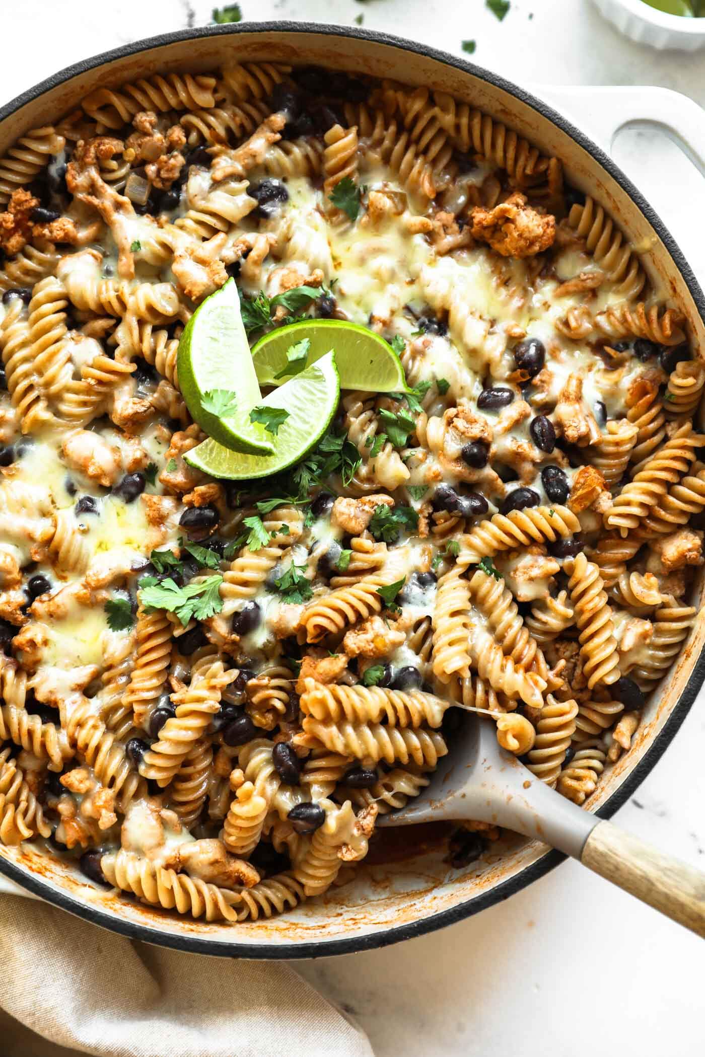 A skillet filled with rotini pasta, black beans, melted cheese, and ground meat, garnished with cilantro and lime wedges, with a serving spoon in the pan.