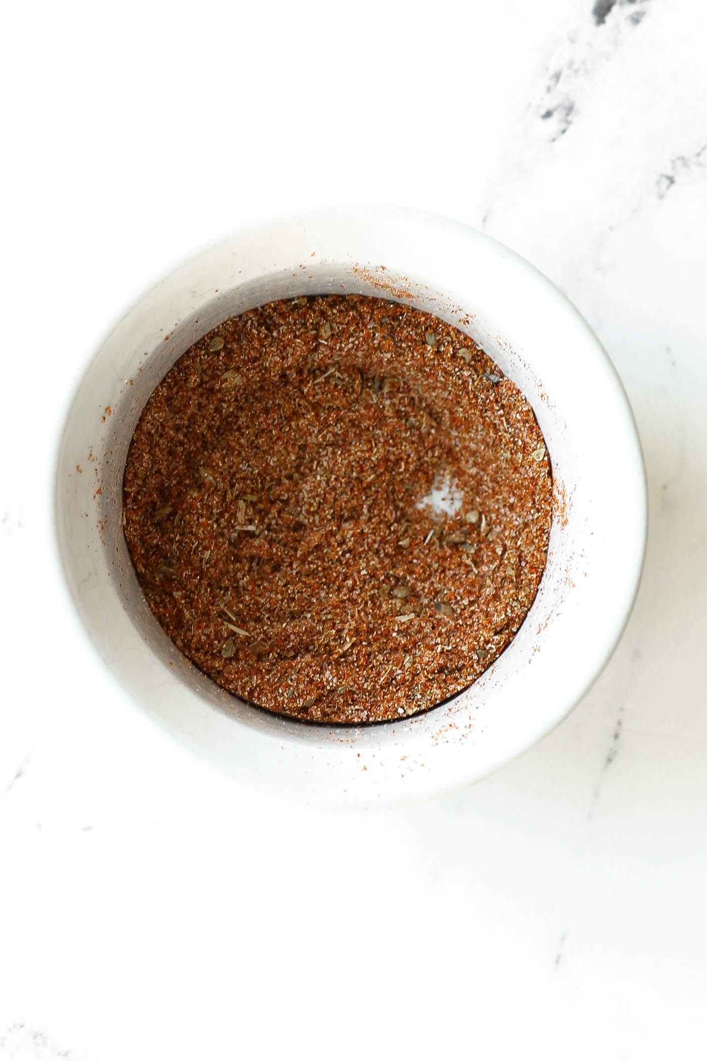 A white bowl filled with a finely ground brown spice mixture sits on a white marble surface.