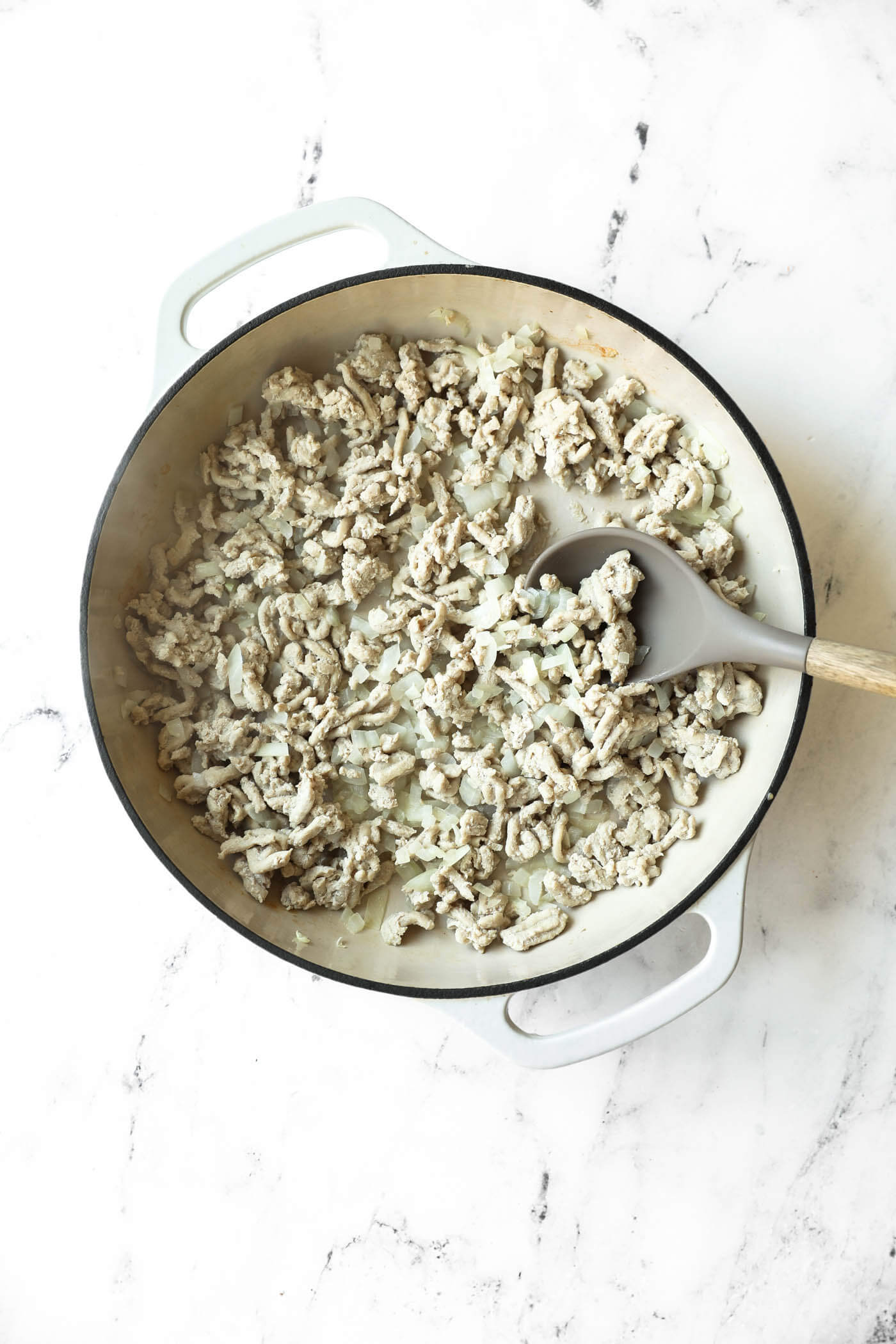 Cooked ground turkey and chopped onions in a white Dutch oven, being stirred with a gray spoon on a white marble surface.