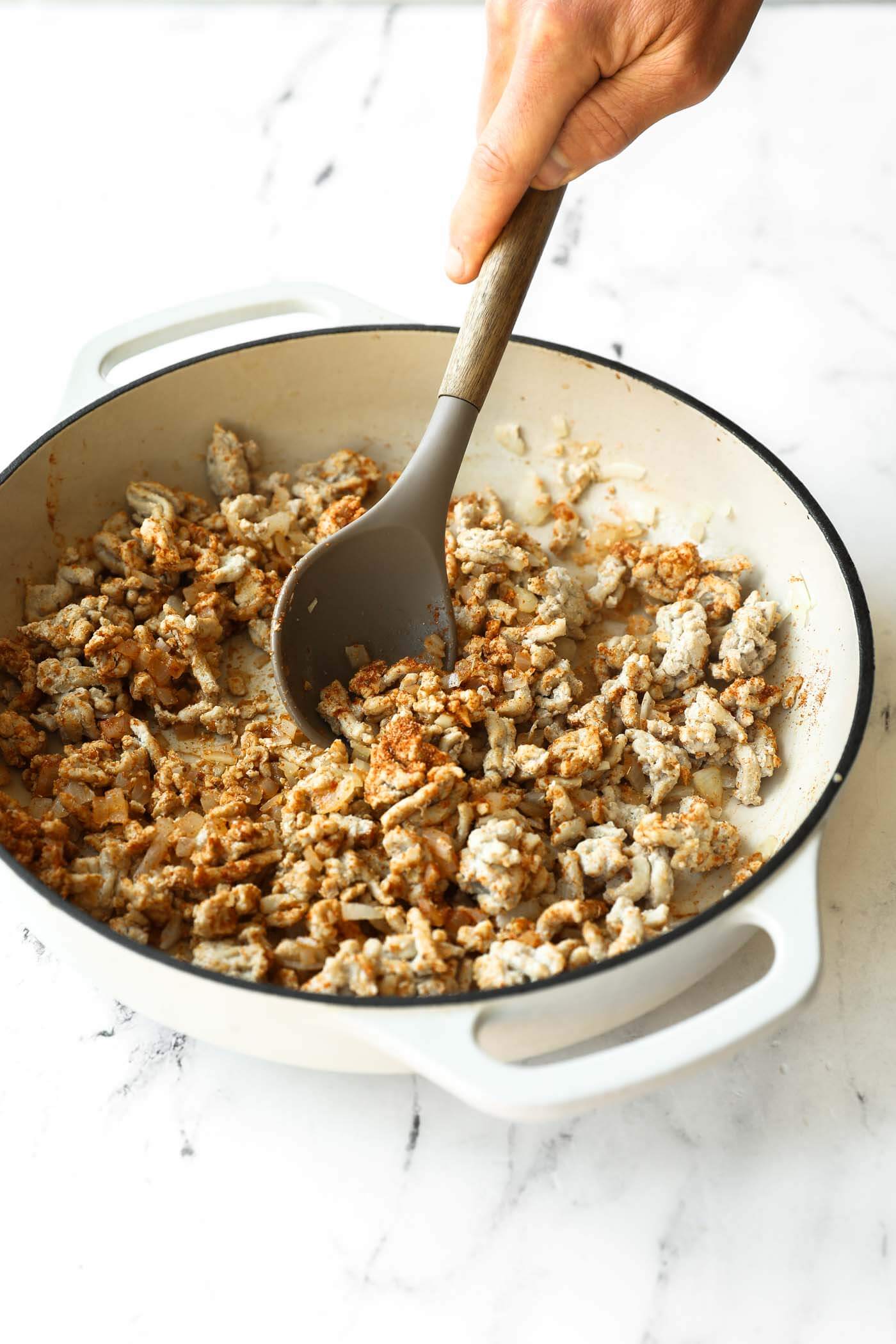 A hand stirs browned ground meat with a gray spatula in a white enameled pot on a marble countertop.