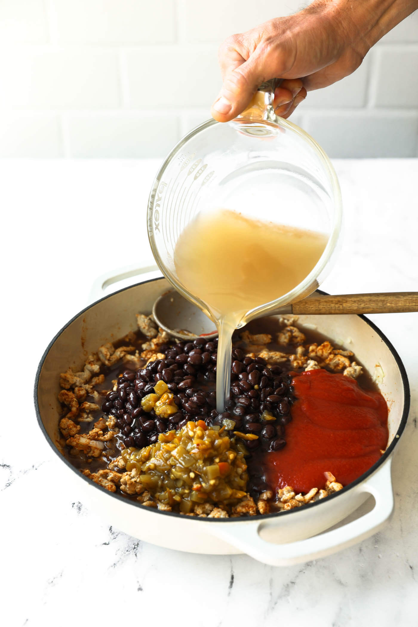 A hand pours broth from a glass measuring cup into a pot containing black beans, diced green chilies, tomato sauce, and browned ground meat on a white countertop.
