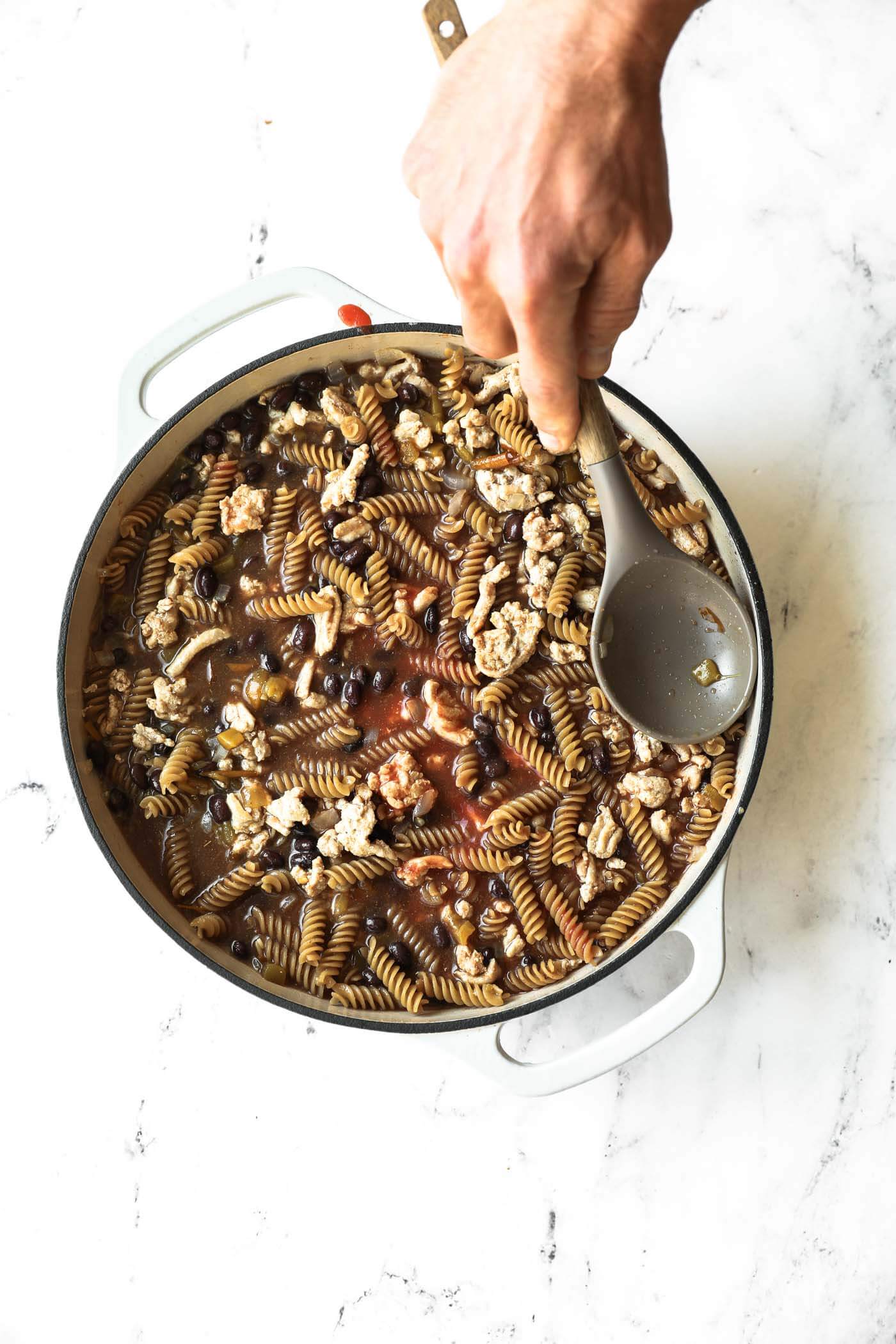 A hand holding a spoon over a pot filled with rotini pasta, ground meat, black beans, and sauce on a white countertop.