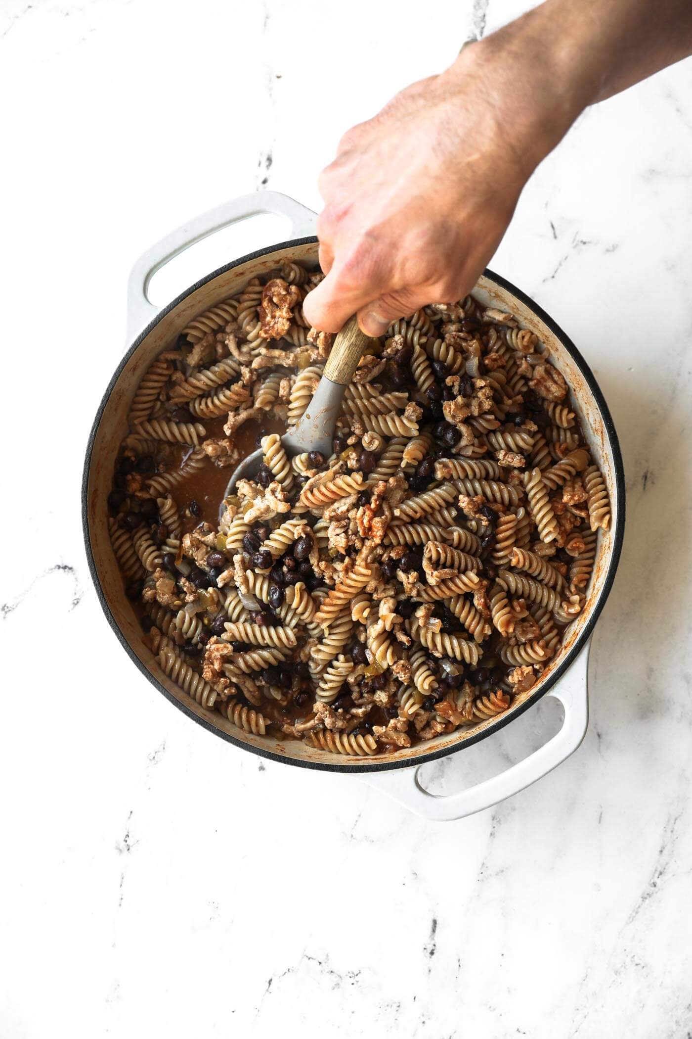 A hand stirs cooked rotini pasta with beans and sauce in a large white pot on a marble surface.
