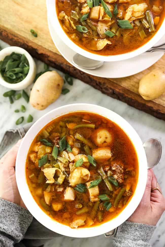 Hamburger soup image overhead picture with two bowls and potatoes and green onions. Hands holding one bowl.