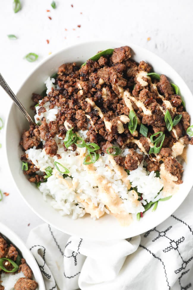 Close up of one bowl of korean beef over rice and greens. Topped with chopped green onion and spicy mayo. 