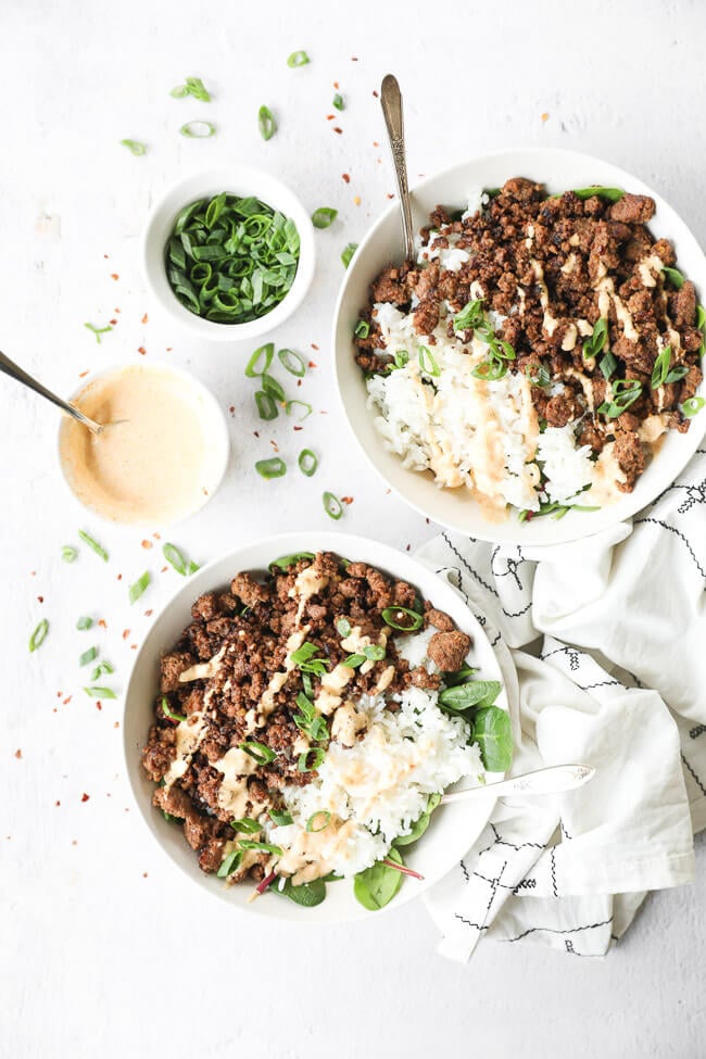 Two bowls of korean beef with rice and greens, topped with chopped green onion and a spicy mayo sauce. Extra sauce and green onion on the side. 