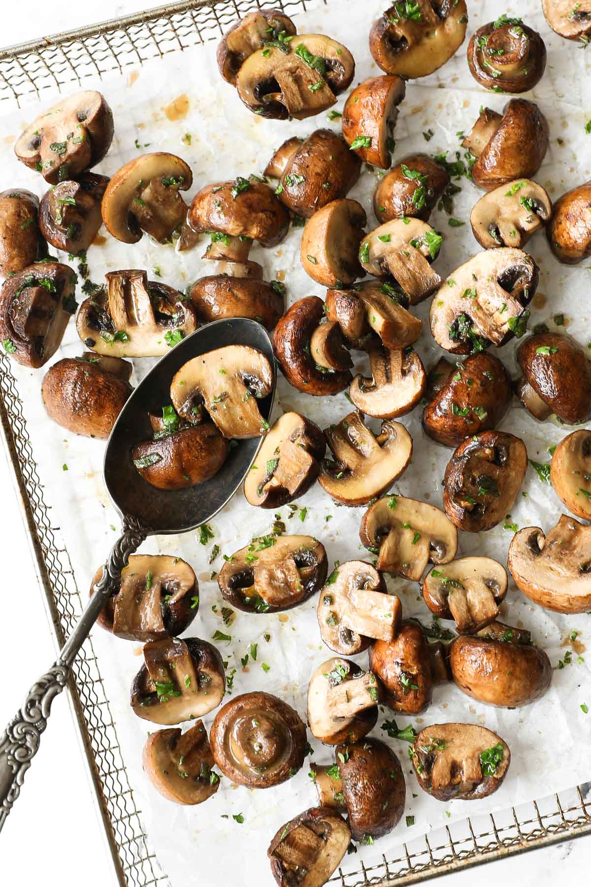 Vertical close up overhead image of air fryer mushrooms in basket with a serving spoon scooping up a couple mushrooms.