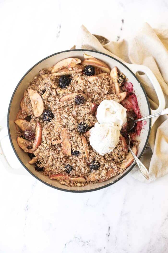 Overhead image of a skillet filled with blackberry and apple crumble. It's being served with two scoops of dairy free ice cream and two spoons.