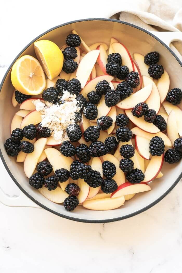 Overhead image of skillet full of apple and blackberry crumble filling ingredients, including apple slices, blackberries, tapioca starch, maple syrup and 2 lemon halves. All before the ingredients have been mixed together.
