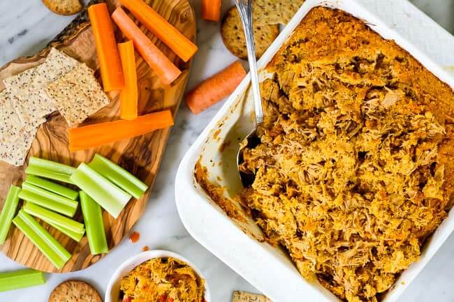 Healthy buffalo chicken dip in baking dish with spoon, crackers, carrots and celery. 