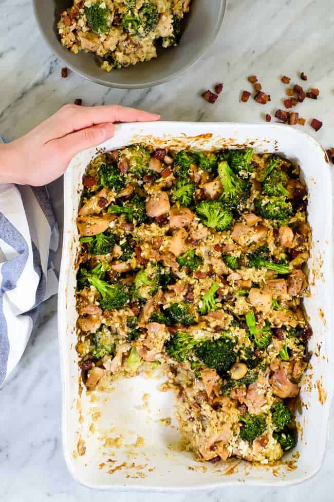 Holding this healthy chicken and broccoli casserole with bacon and cauliflower rice in a creamy sauce in a casserole dish. 