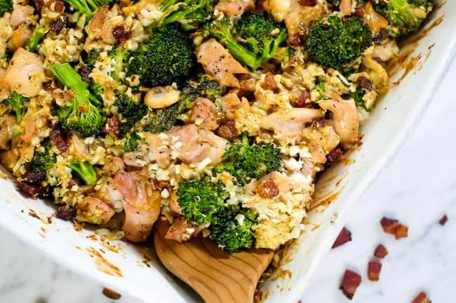 Close up of this healthy chicken and broccoli casserole with bacon and cauliflower rice in a creamy sauce in a casserole dish with a serving spoon. 