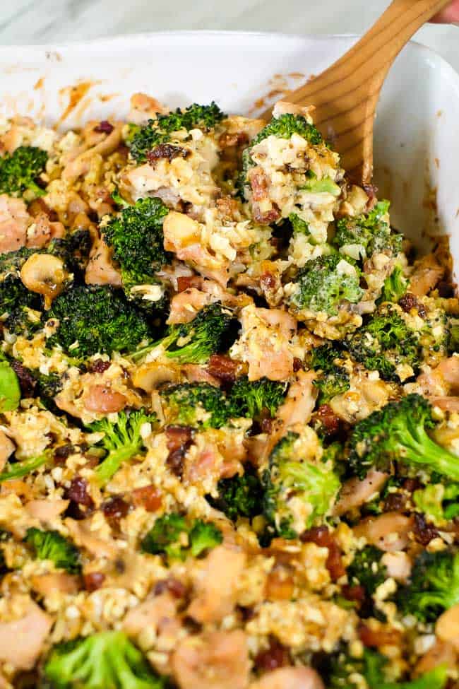 Close up of this healthy chicken and broccoli casserole with bacon and cauliflower rice in a creamy sauce in a casserole dish with a serving spoon. 