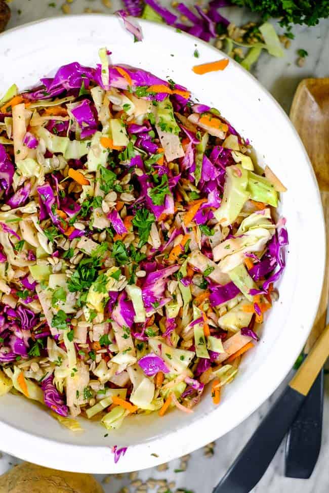 Healthy coleslaw in a serving bowl with serving utensils on table along with slaw ingredients. 