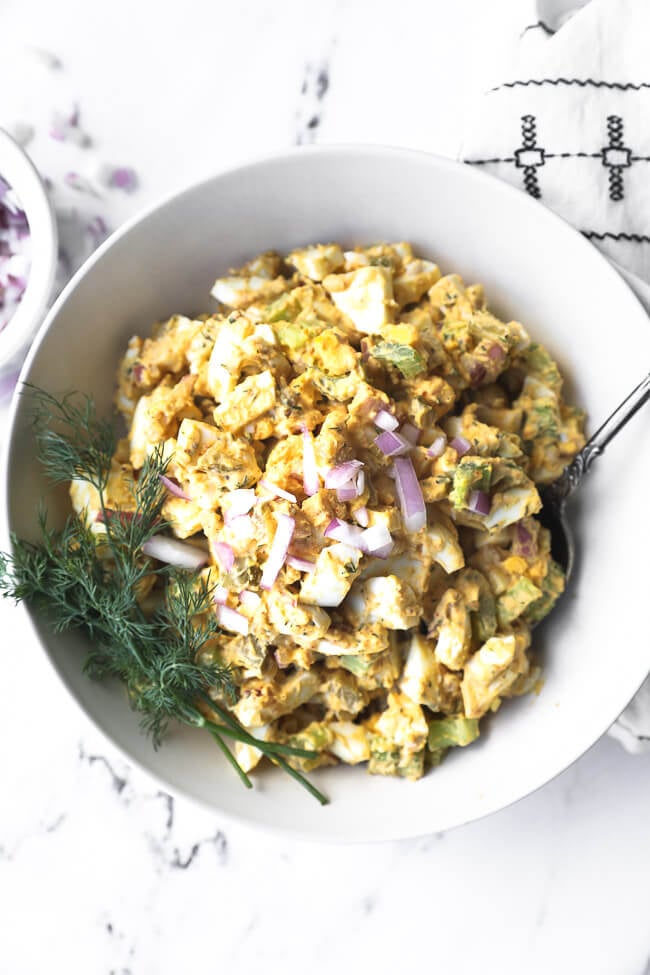 Overhead image of healthy egg salad in a bowl with a serving spoon. Topped with chopped red onion and fresh dill. 