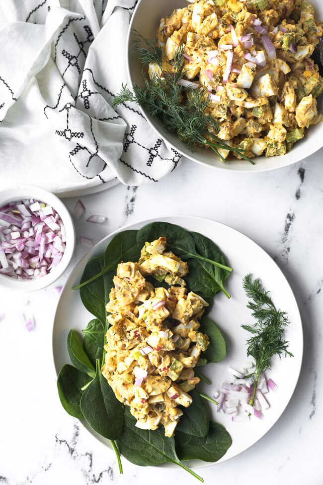 Healthy egg salad served over a bed of greens on a plate. Chopped red onion on the side and extra egg salad in a serving bowl on the side. 