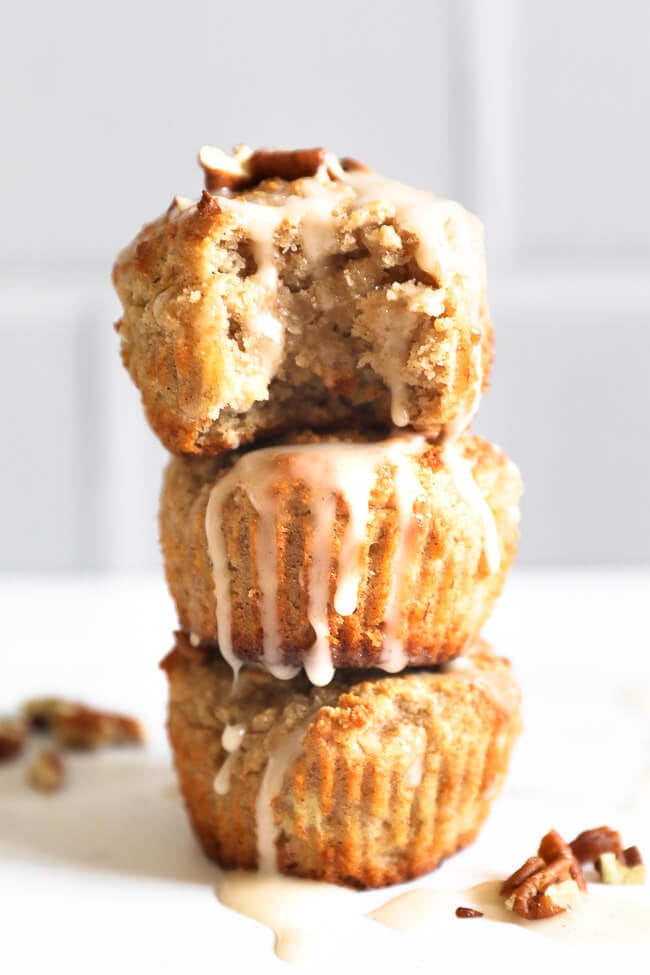 Three banana muffins stacked with the top muffin having a bite taken out. Drizzled with glaze and topped with chopped pecans. 