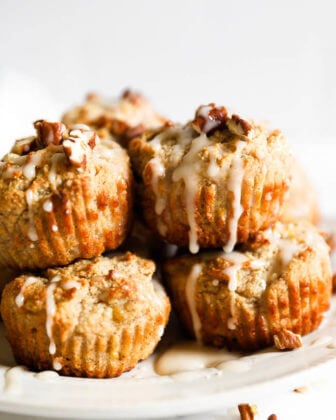 Gluten free and vegan banana muffins stacked on a plate with maple glaze drizzled on top and sprinkled with chopped pecans.