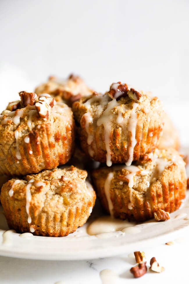 Vegan banana muffins stacked up on a plate with maple glaze and chopped pecans sprinkled on top. 