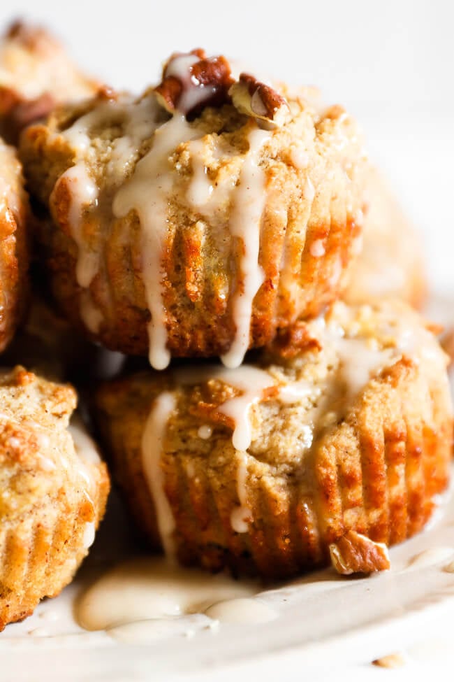 Close up of vegan, gluten free banana muffins on plate. Drizzle dripping down the side of the muffin and chopped pecans on top. 