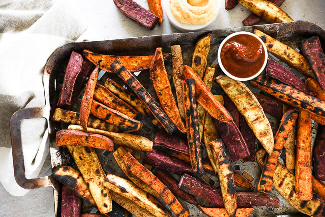 Horizontal overhead image of grilled sweet potato fries on a grilling pan with ketchup and chipotle aioli on the side. 