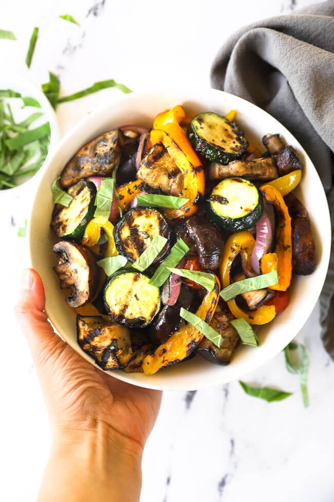 Holding a bowl of grilled veggies tossed in a marinade with chopped basil on top. 
