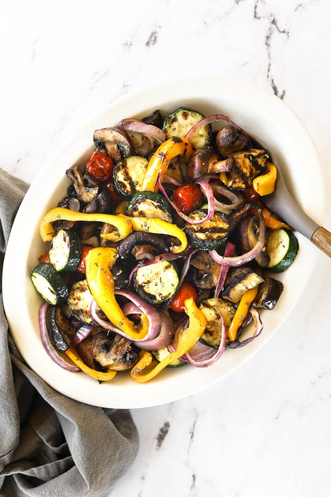 Overhead image of an oval bowl full of grilled veggies tossed in a marinade with a serving spoon.