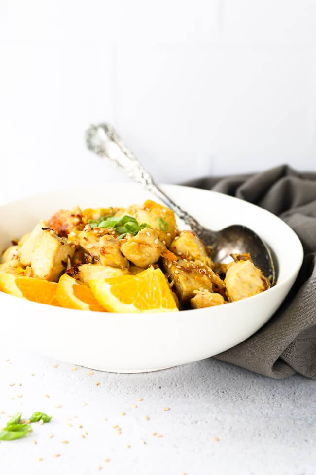 Straight on shot of orange chicken in a bowl with orange wedges and a spoon