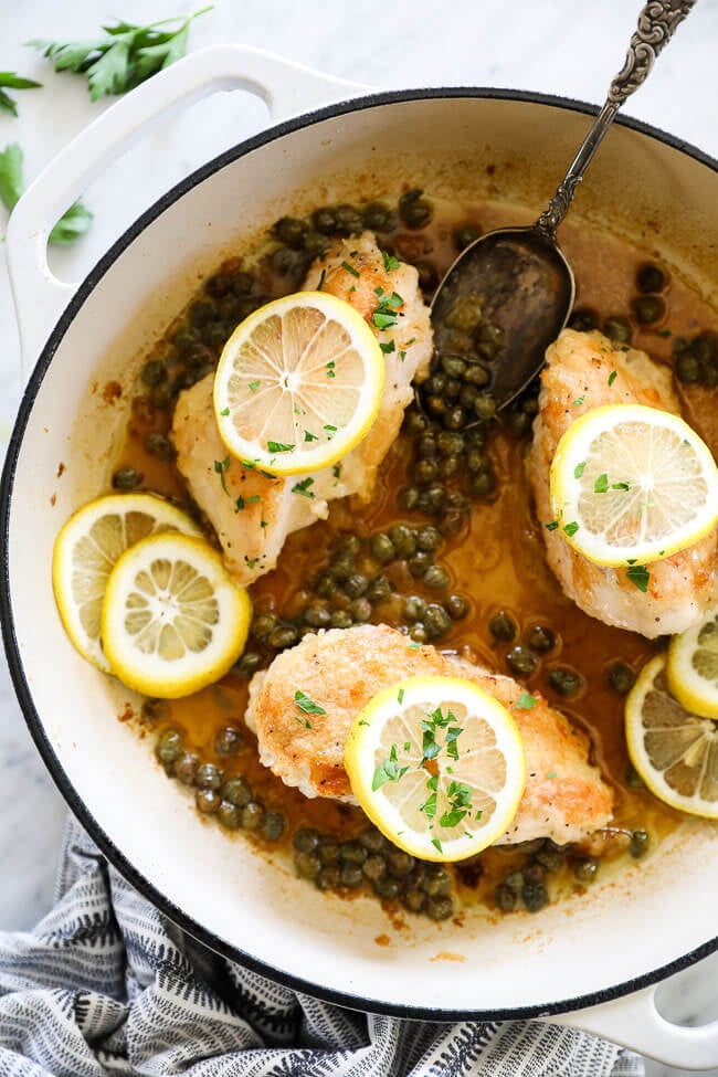 Vertical overhead image of healthy lemon chicken piccata in a large skillet with sauce, capers, lemon rounds and chopped parsley. Serving spoon in skillet. 