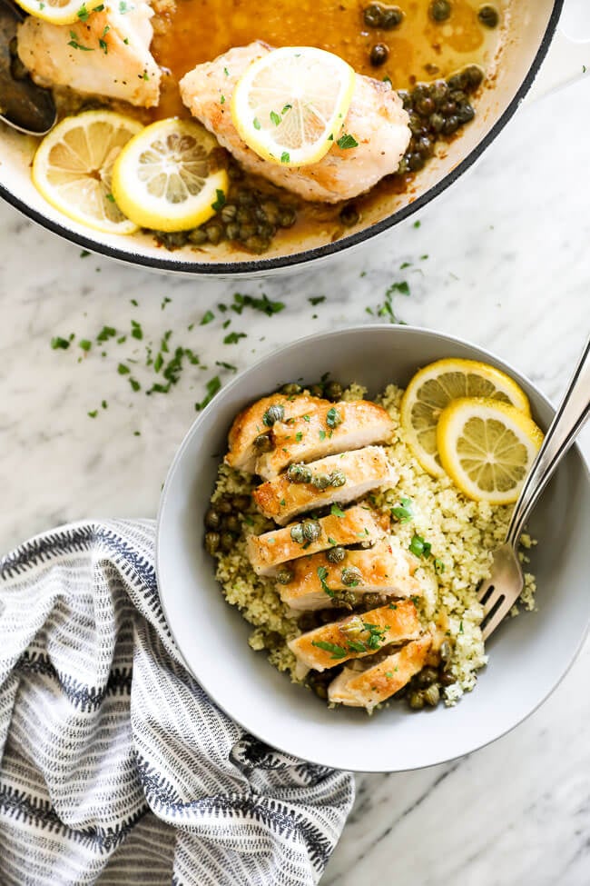 Vertical overhead image of healthy lemon chicken piccata sliced and served in a bowl with lemon rounds and chopped parsley. Fork dug into bowl. 