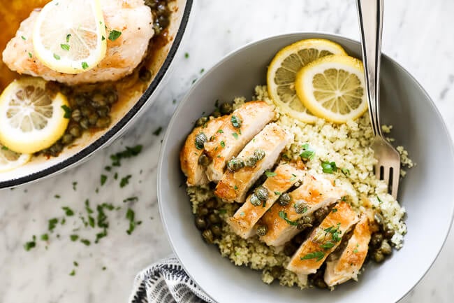 Horizontal overhead image of healthy lemon chicken piccata sliced and served in a bowl with cauliflower rice and lemon rounds. Fork dug into bowl. 