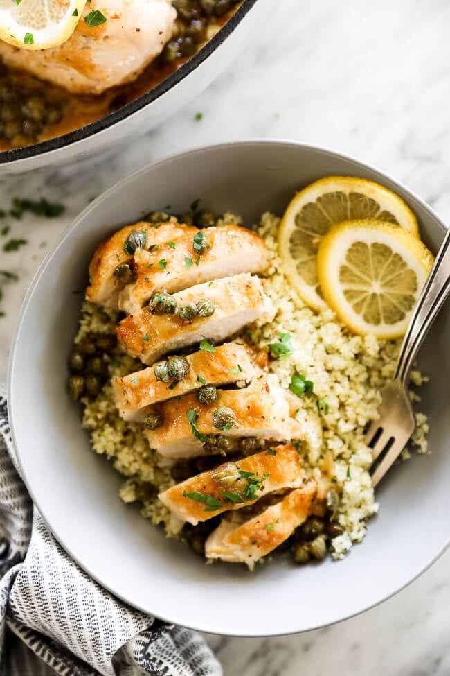 Vertical overhead close up image of healthy lemon chicken piccata in a bowl served over cauliflower rice with lemon rounds and chopped parsley. Fork dug into bowl. 