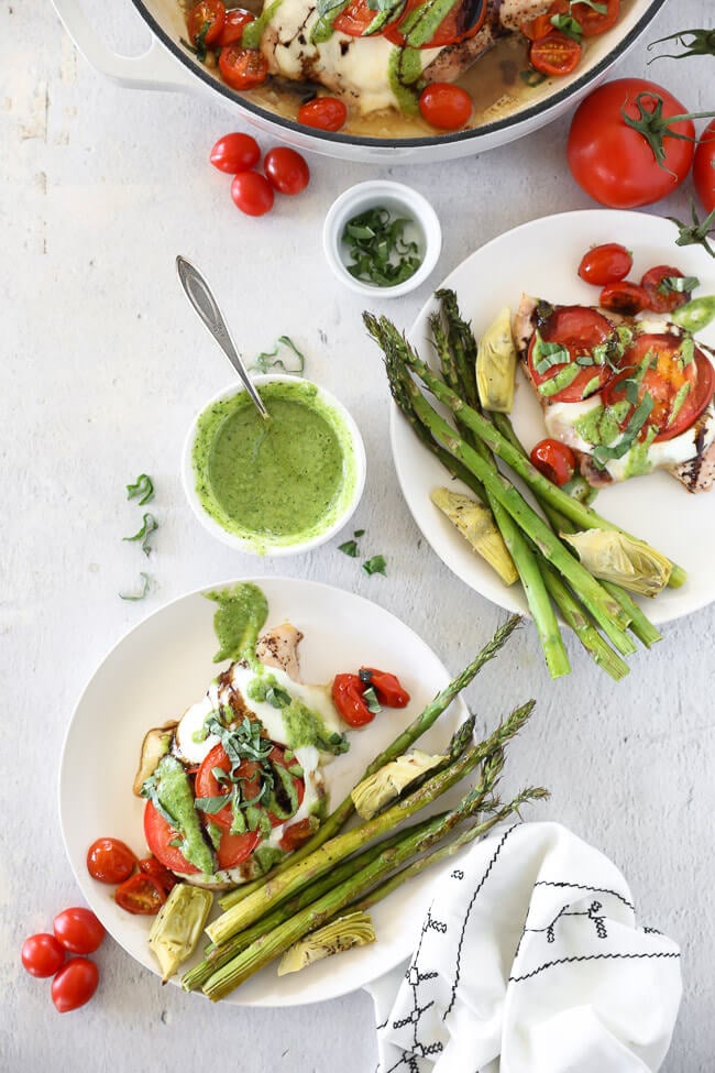 Vertical overhead image of chicken caprese served on two plates with a side of asparagus and artichoke hearts. Chicken breasts topped with mozzarella cheese, tomatoes, pesto, balsamic and fresh basil. 
