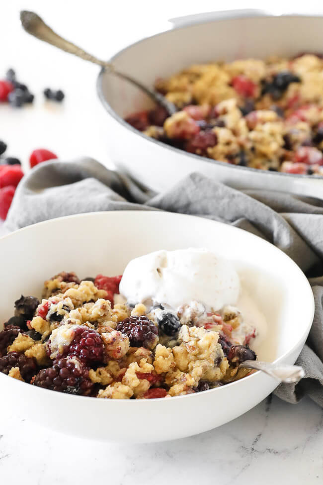 Angled vertical image of berry crisp served in a bowl with a scoop of ice cream on top and a spoon dug in. 