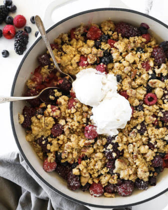 Overhead vertical image of mixed berry crisp in skillet with two spoons dug in and two scoops of ice cream on top.