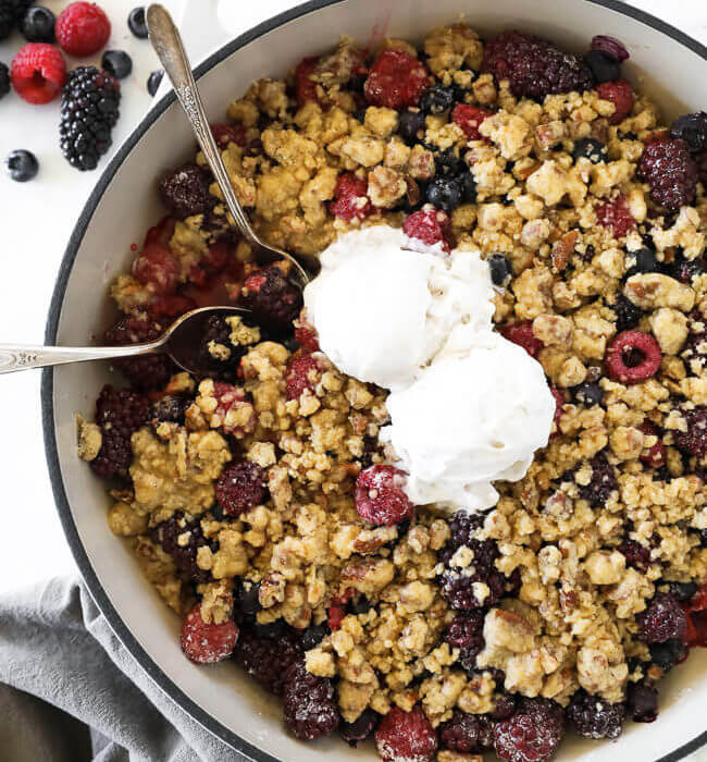 Overhead vertical image of mixed berry crisp in skillet with two spoons dug in and two scoops of ice cream on top.