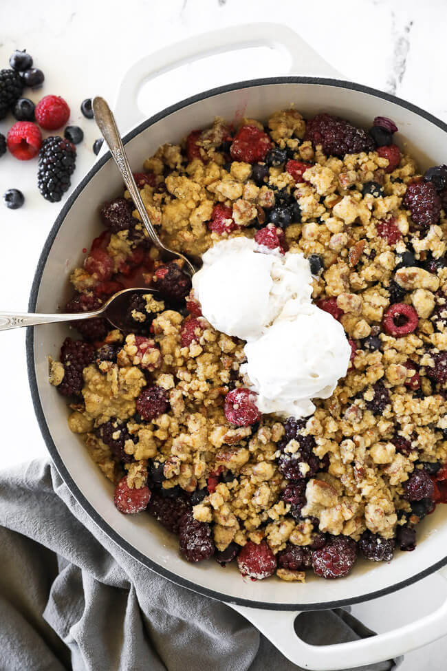 Overhead vertical image of mixed berry crisp in skillet with two spoons dug in and two scoops of ice cream on top.