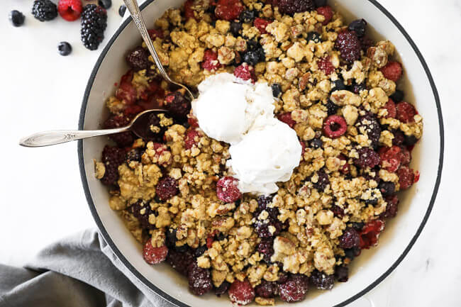 Overhead horiztonal image of mixed berry crisp in skillet with two spoons dug in and two scaoops of ice cream on top. 