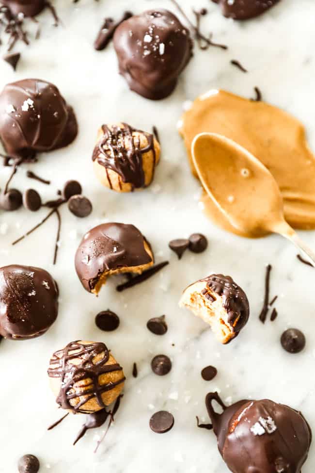 peanut butter balls, some with chocolate drizzle and some coated in chocolate spread out on marble and sprinkled with flaky sea salt with a spoonful of peanut butter. 