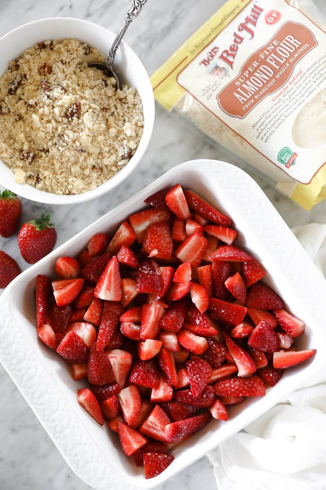 Vertical overhead image of strawberries in a baking dish with topping in a small bowl on the side and bob's red mill almond flour. 